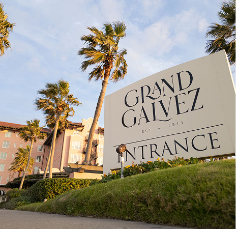 Grand Galvez entrance sign with palm trees and the pink hotel behind it